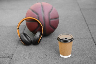 Photo of Paper cup of hot coffee, basketball and headphones on floor outdoors. Takeaway drink