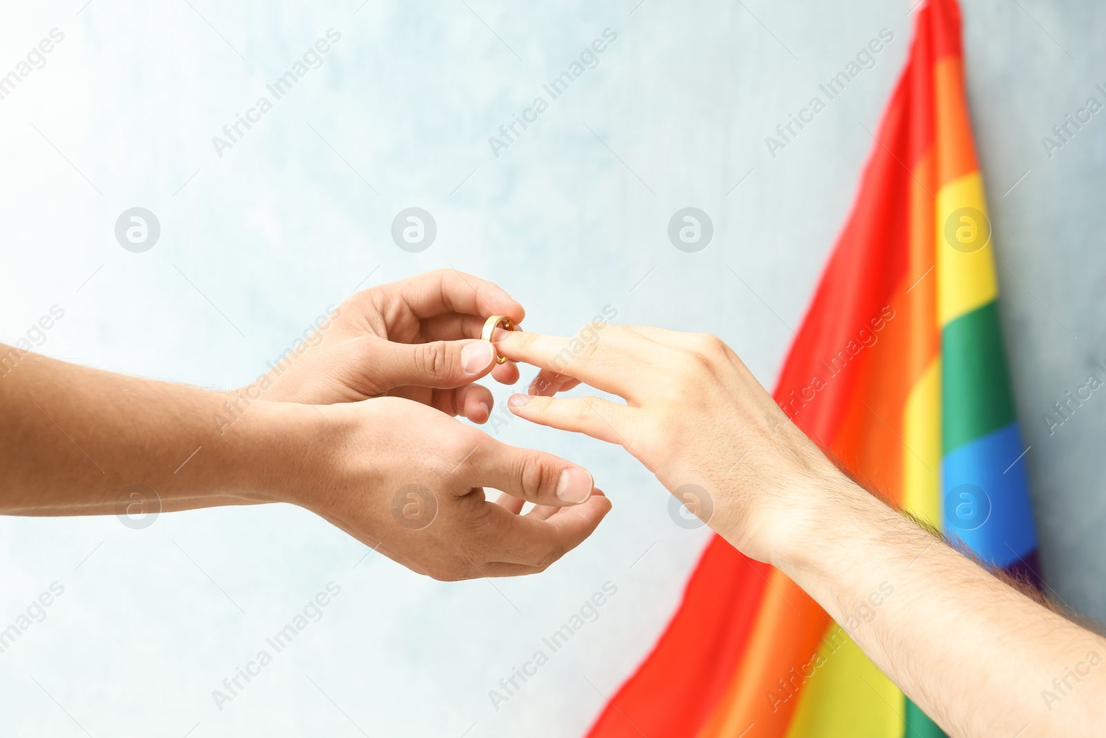 Photo of Young man putting wedding ring on his boyfriend's finger near rainbow flag. Gay marriage