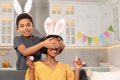 Photo of Cute African American son covering his mother's eyes with hands while she holding Easter eggs in kitchen