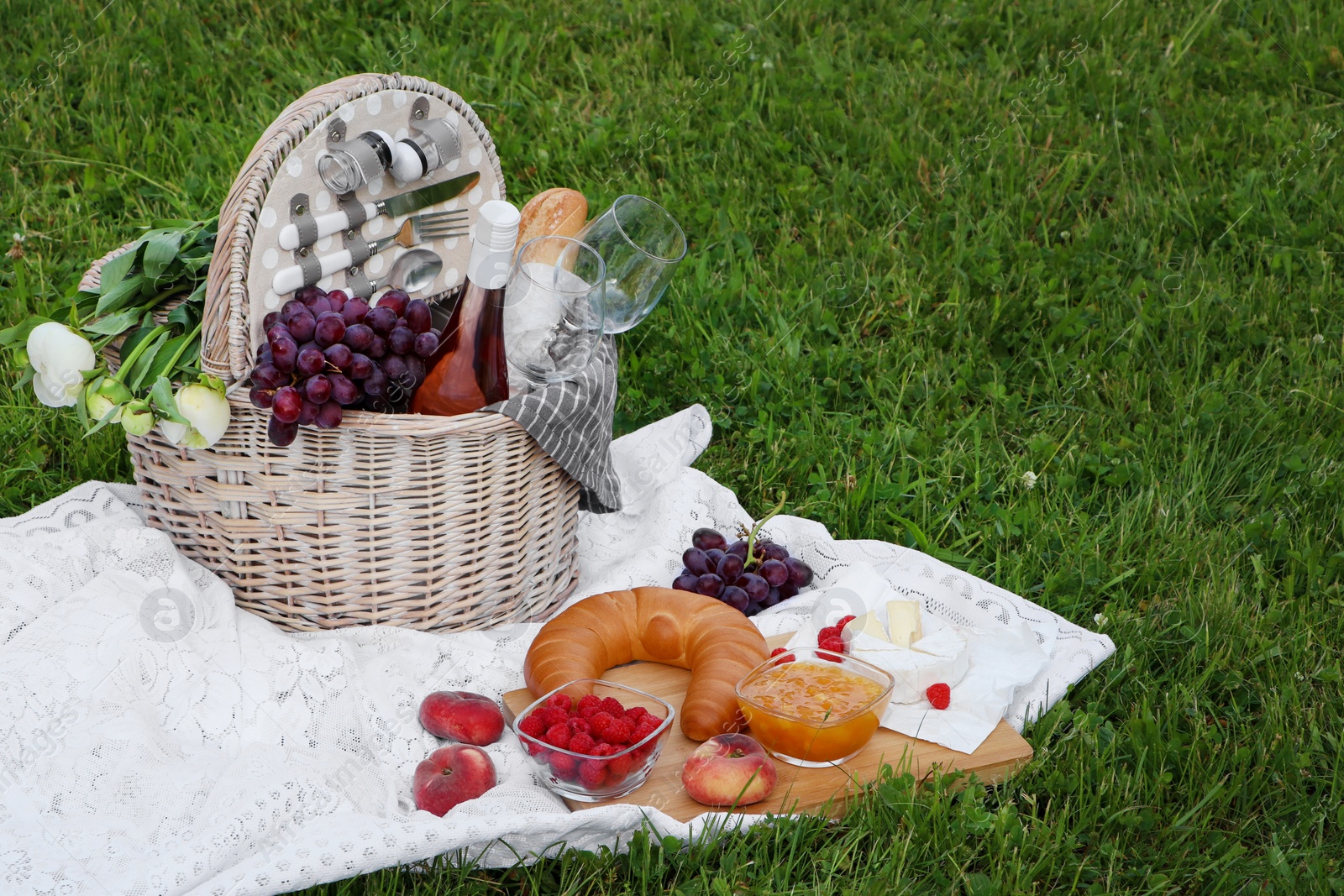 Photo of Picnic blanket with tasty food, flowers, basket and cider on green grass outdoors