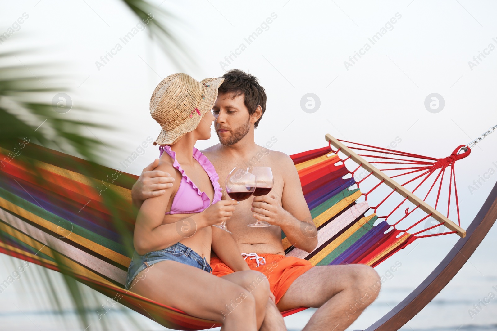 Photo of Young couple resting with glasses of wine in hammock on beach