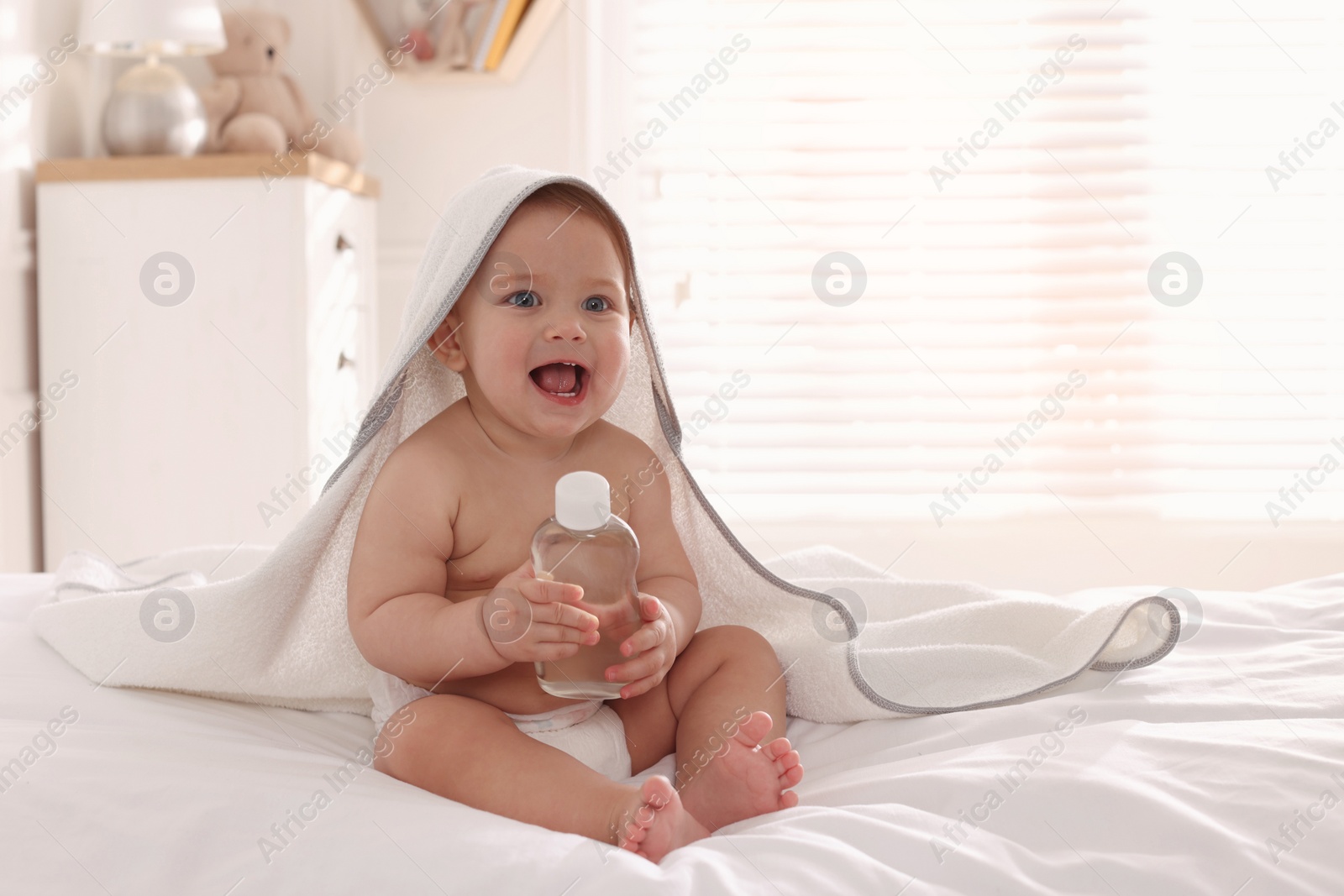 Photo of Cute little baby with towel and bottle of massage oil on bed at home