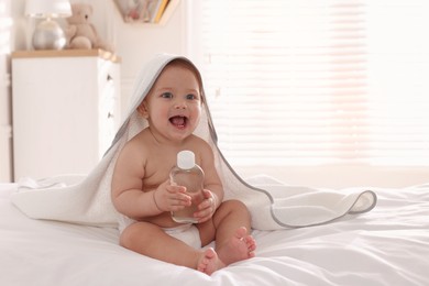 Cute little baby with towel and bottle of massage oil on bed at home
