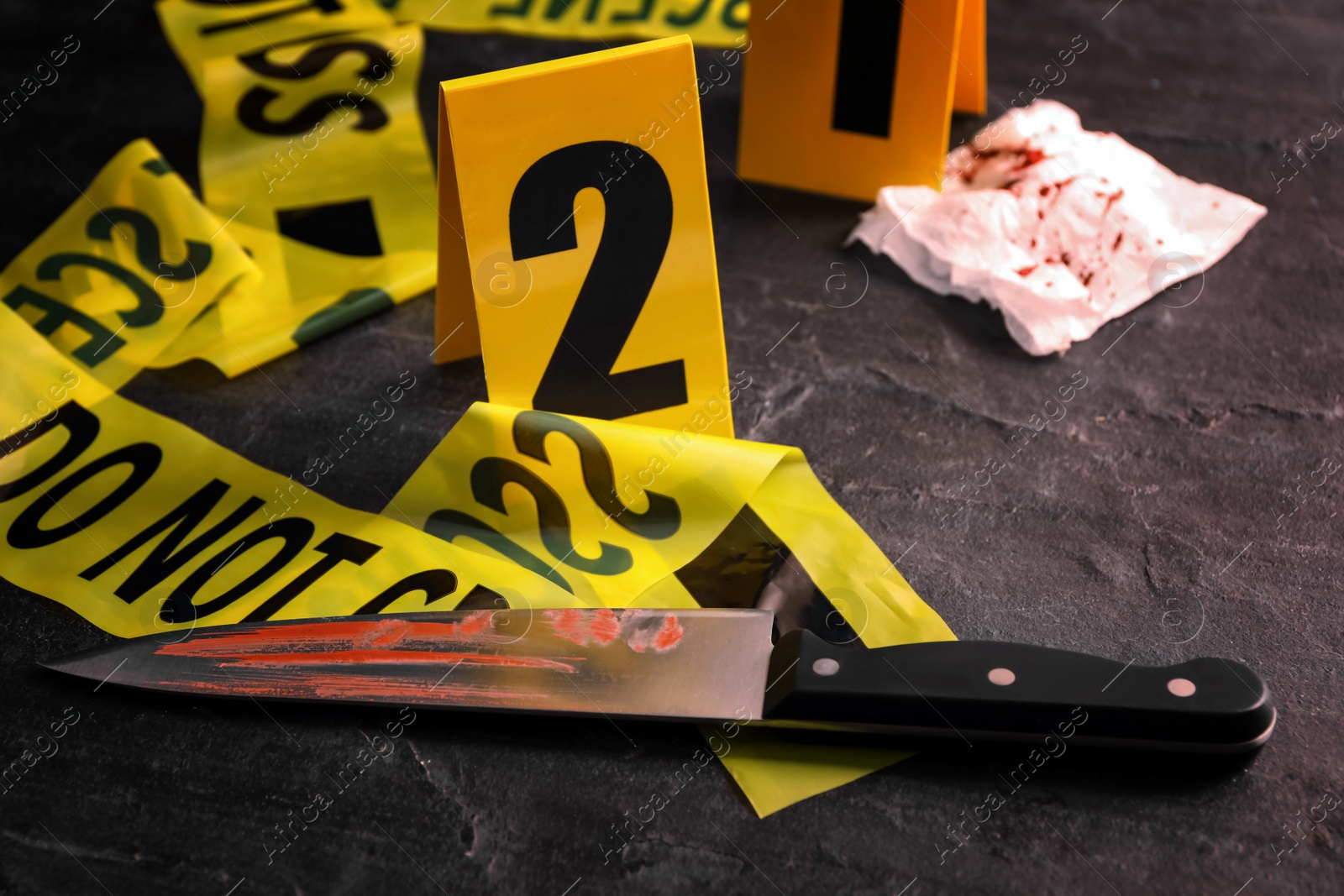 Photo of Bloody knife, yellow tape and evidence marker on black slate table, closeup. Crime scene