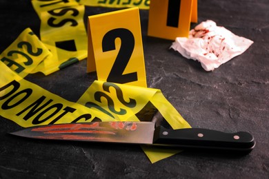 Bloody knife, yellow tape and evidence marker on black slate table, closeup. Crime scene