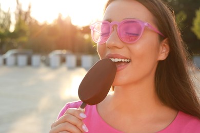 Photo of Beautiful young woman eating ice cream glazed in chocolate on city street, closeup