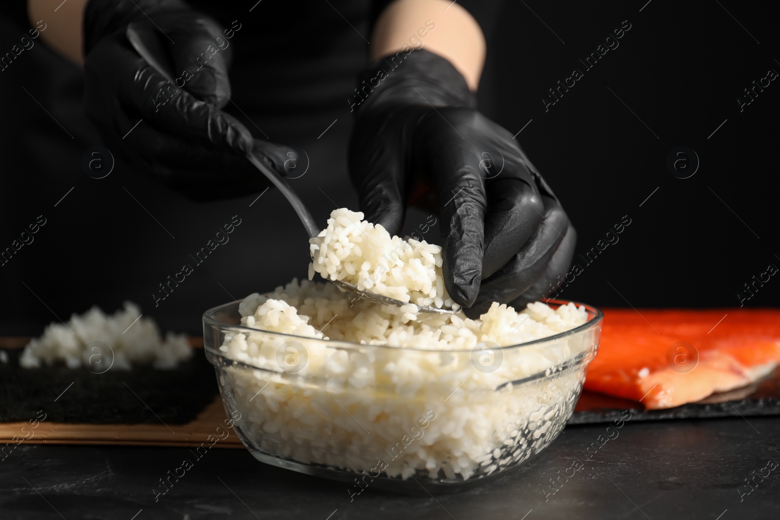 Photo of Chef in gloves taking cooked rice for sushi with spoon at dark table, closeup