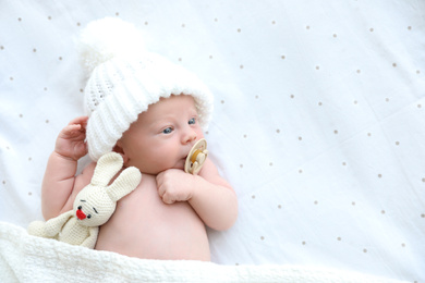 Cute newborn baby in white knitted hat with toy lying on bed, top view. Space for text