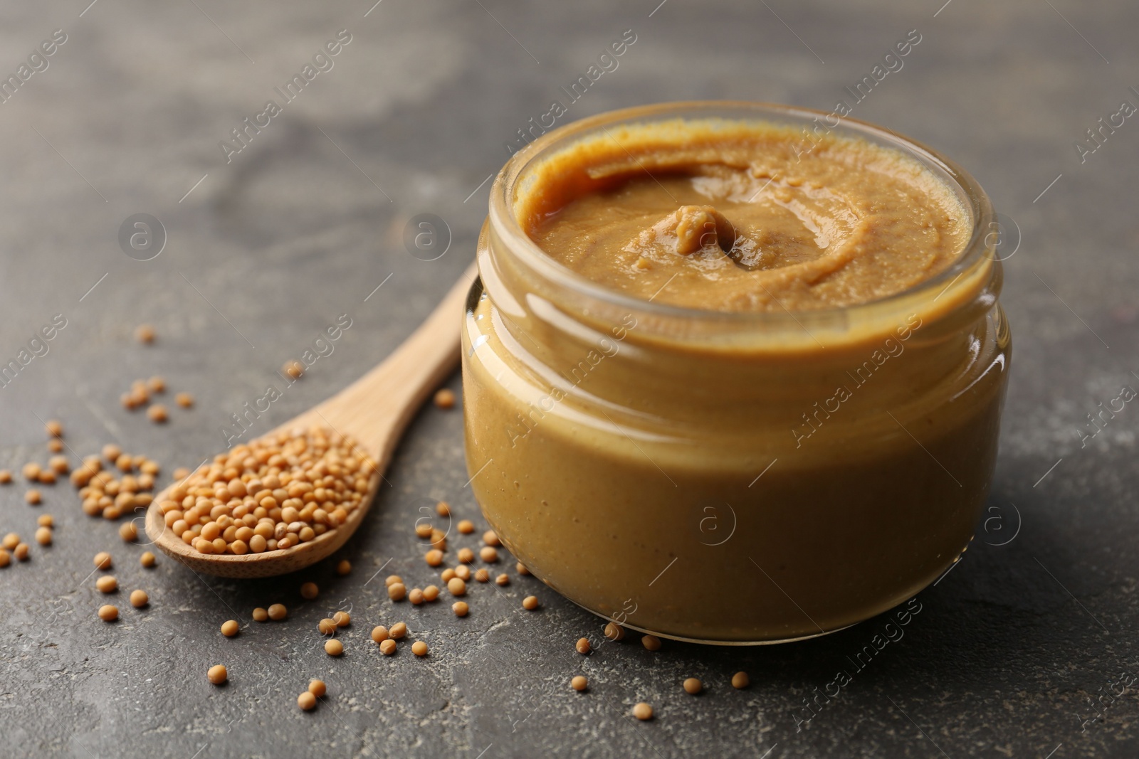 Photo of Tasty mustard sauce in jar and spoon with dry seeds on grey textured table, closeup