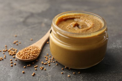 Tasty mustard sauce in jar and spoon with dry seeds on grey textured table, closeup