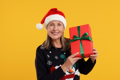 Photo of Happy senior woman in Christmas sweater and Santa hat holding gift on orange background
