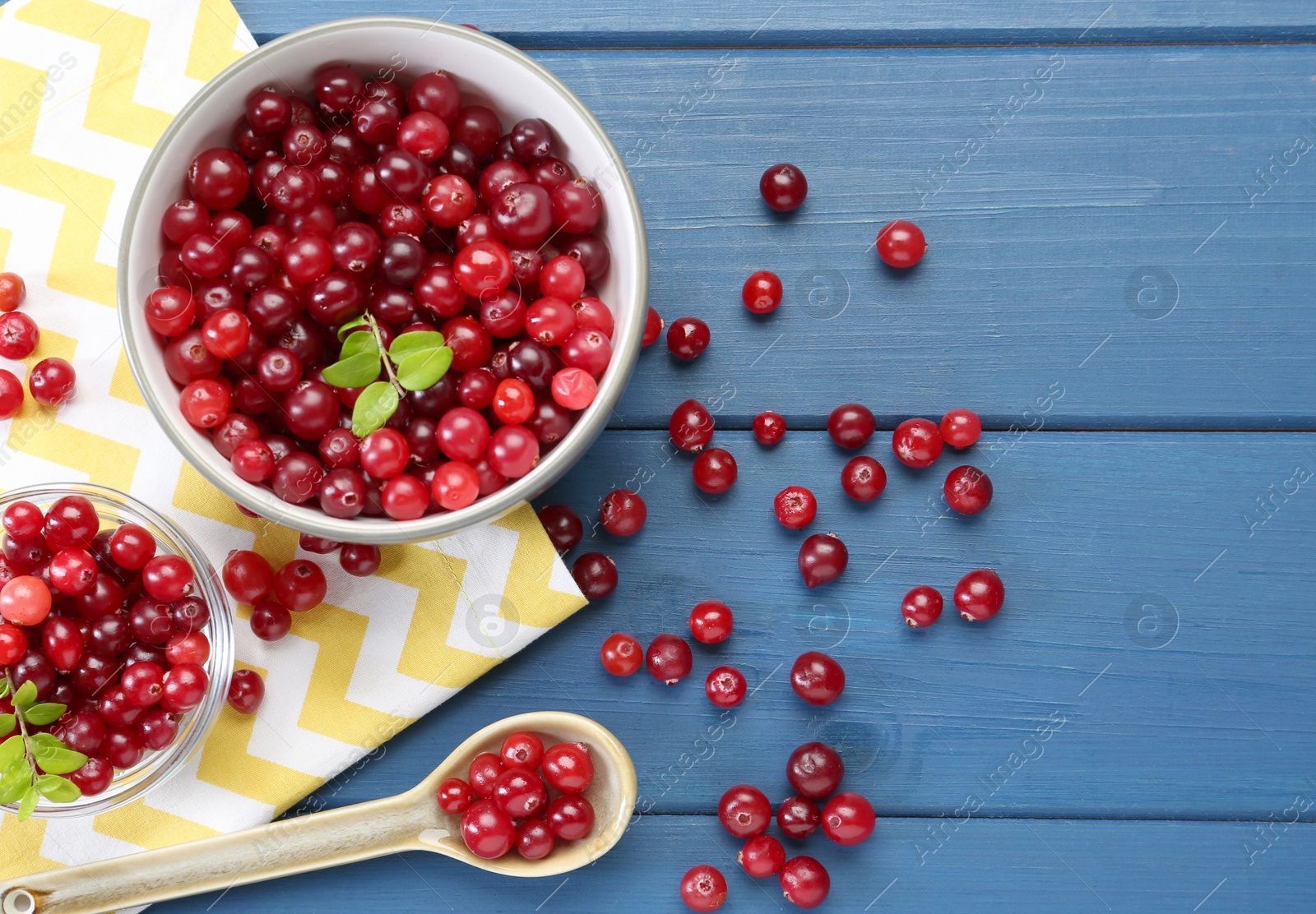 Photo of Fresh ripe cranberries in bowls and spoon on blue wooden table, flat lay