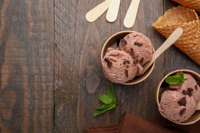 Photo of Paper cups with tasty chocolate ice cream, sticks, mint leaves and cones on wooden table, flat lay. Space for text