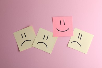 Photo of Choice concept. Sticky note with happy emoticon among beige papers with sad emojis on pink background, top view