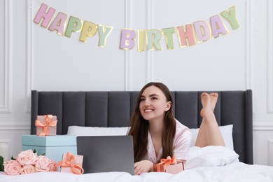 Photo of Beautiful young woman using laptop near gift boxes and rose flowers on bed in room. Happy Birthday
