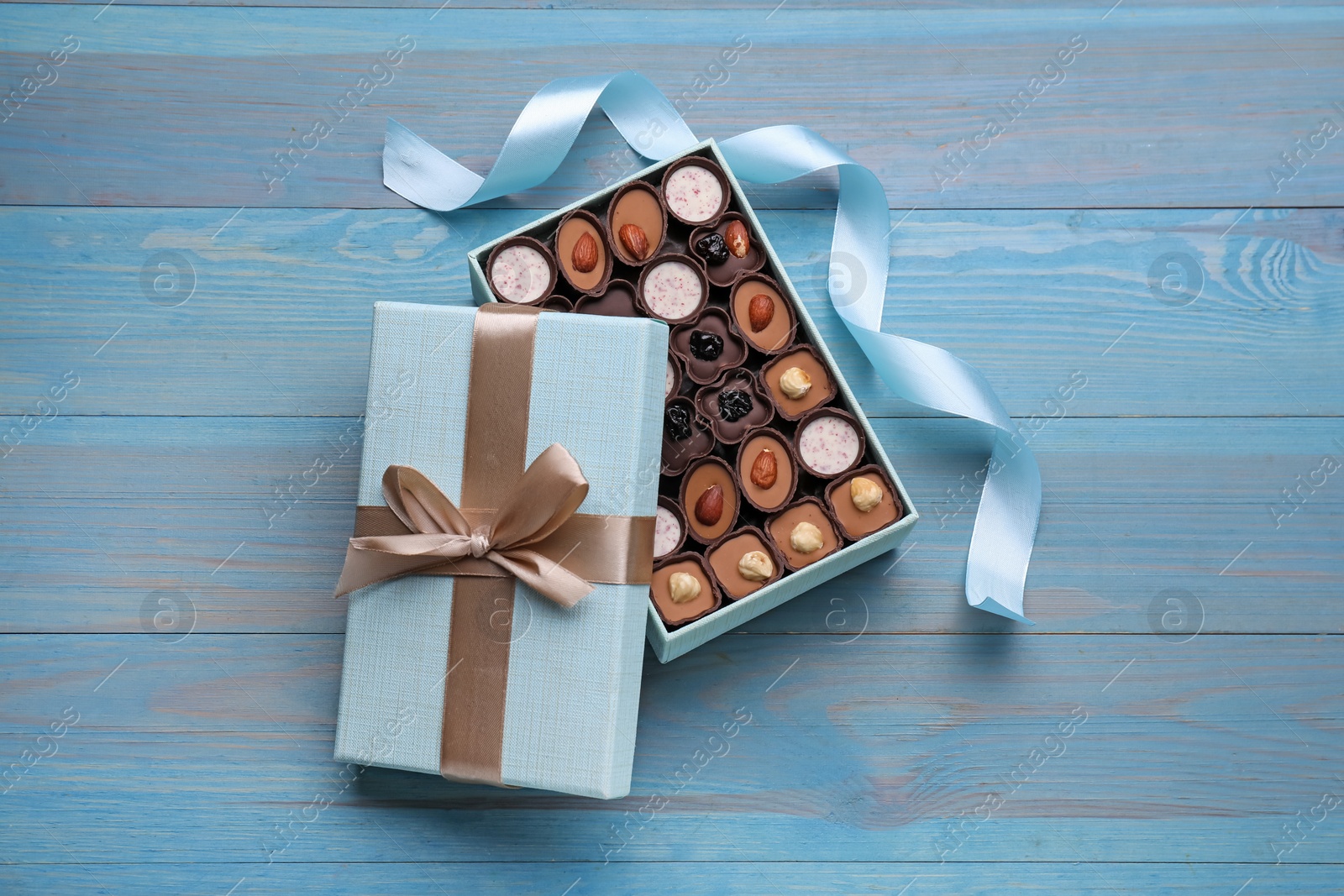 Photo of Open box of delicious chocolate candies and color ribbon on light blue wooden table, flat lay
