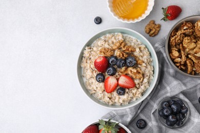 Photo of Tasty oatmeal with strawberries, blueberries and walnuts surrounded by ingredients on grey table, flat lay. Space for text