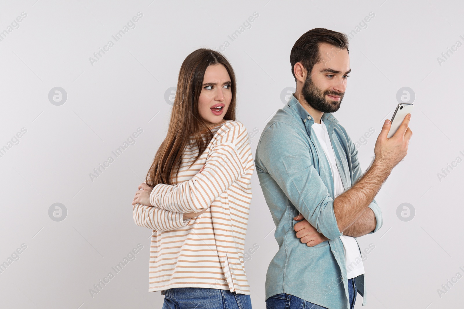 Photo of Indignant young woman looking into boyfriend's smartphone on light grey background. Relationship problems