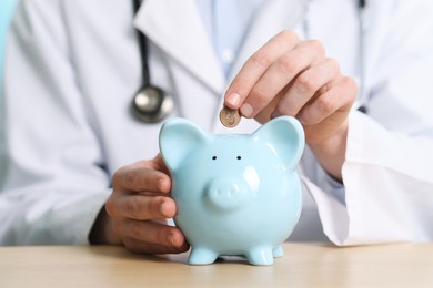 Photo of Doctor putting coin into piggy bank at wooden table, closeup