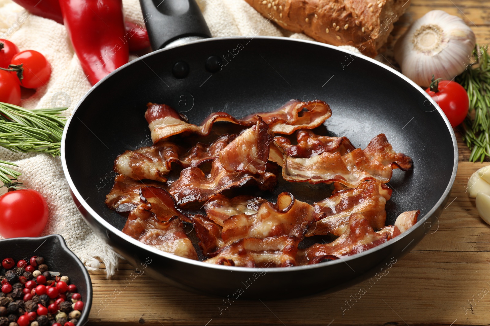 Photo of Delicious bacon slices in frying pan and products on wooden table, closeup