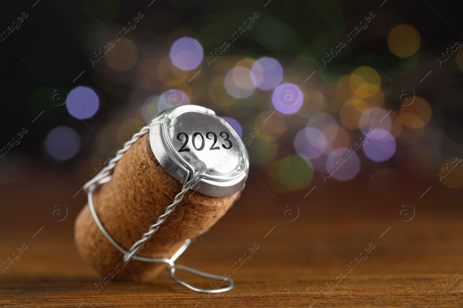Image of Cork of sparkling wine and muselet cap with engraving 2023 on wooden table against blurred festive lights, closeup. Bokeh effect
