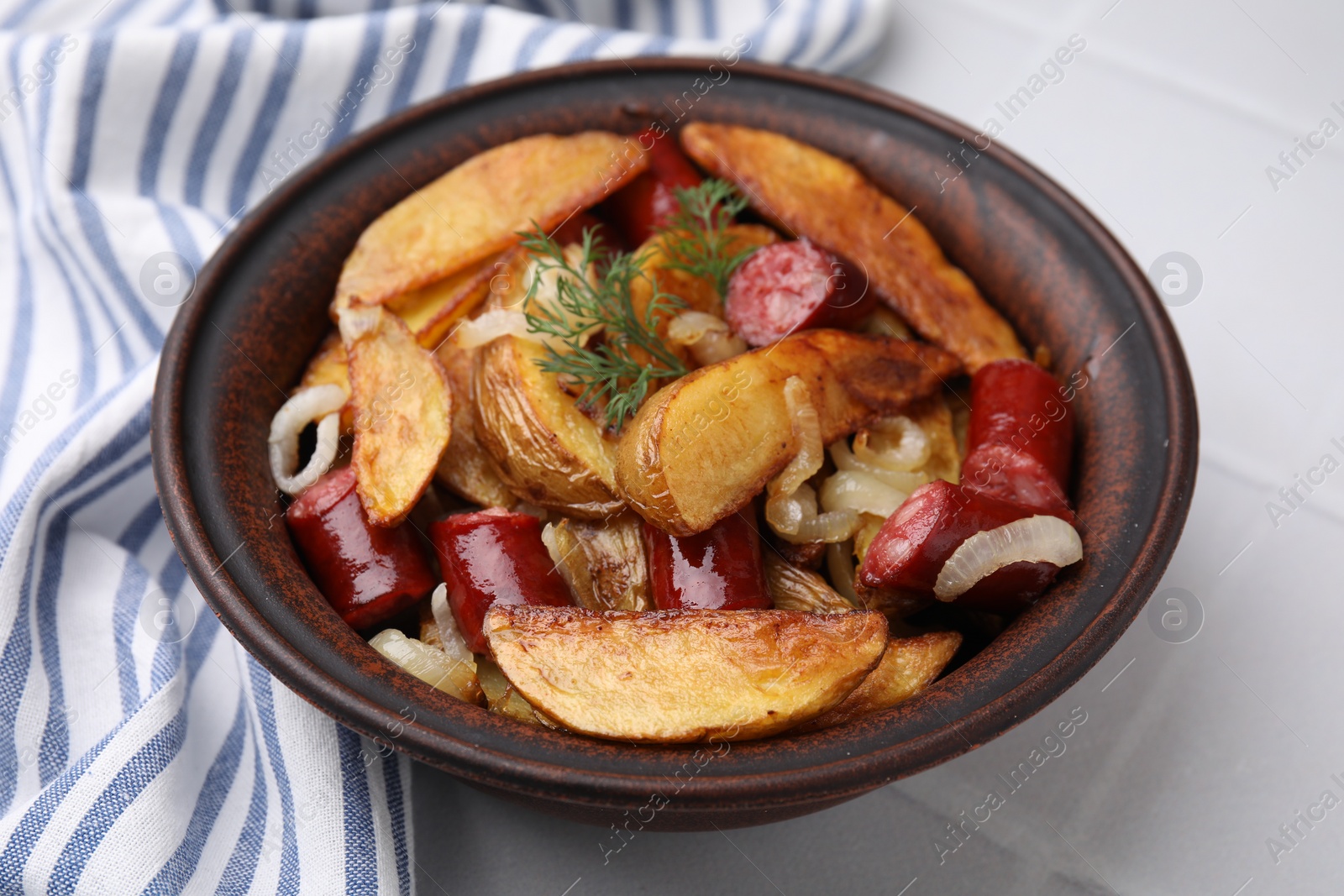 Photo of Delicious baked potato with thin dry smoked sausages, onion and dill in bowl on white table, closeup