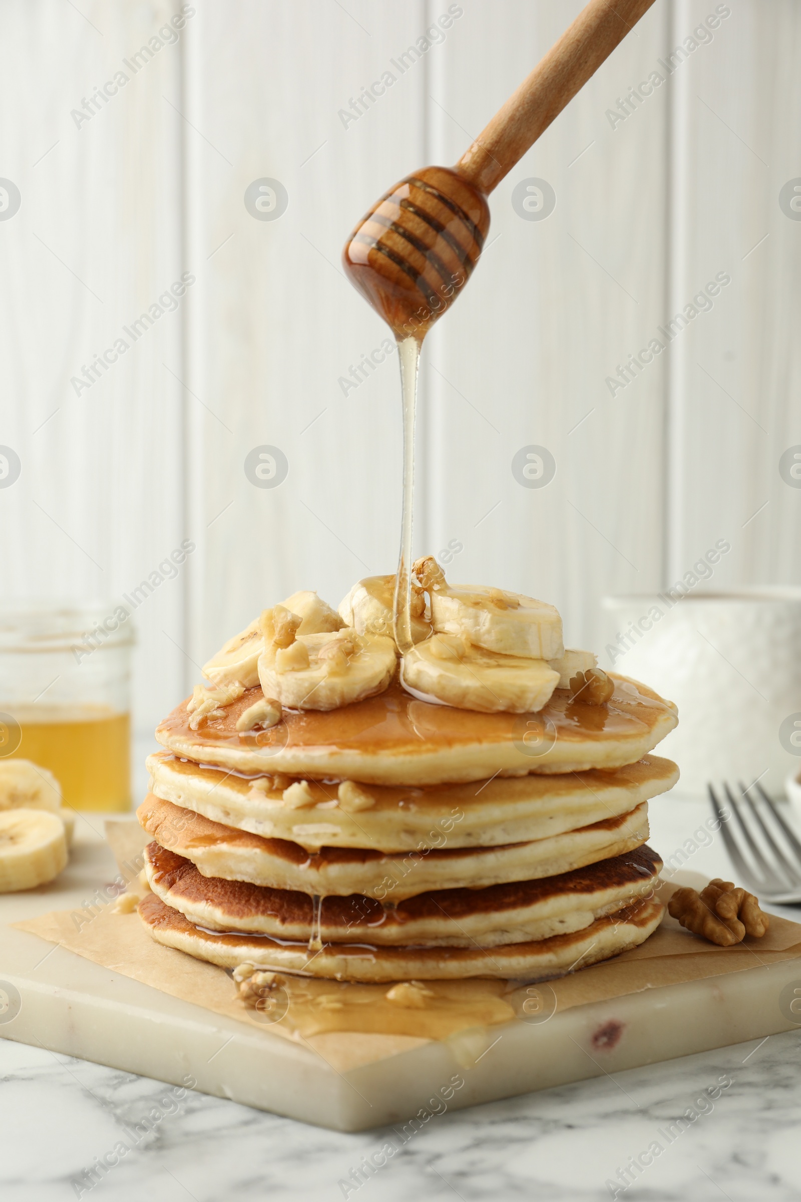 Photo of Pouring honey from dipper onto delicious pancakes with bananas and walnuts at white marble table