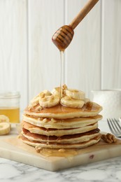 Photo of Pouring honey from dipper onto delicious pancakes with bananas and walnuts at white marble table