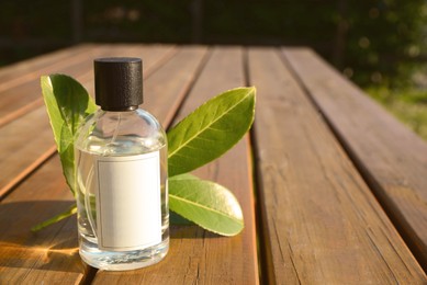 Photo of Bottle of perfume and green leaves on wooden table outdoors, space for text