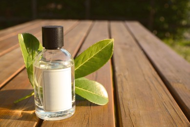 Bottle of perfume and green leaves on wooden table outdoors, space for text