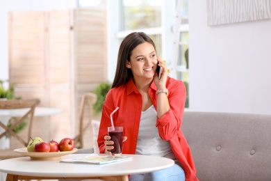 Photo of Young woman using mobile phone while drinking tasty healthy smoothie at table, indoors