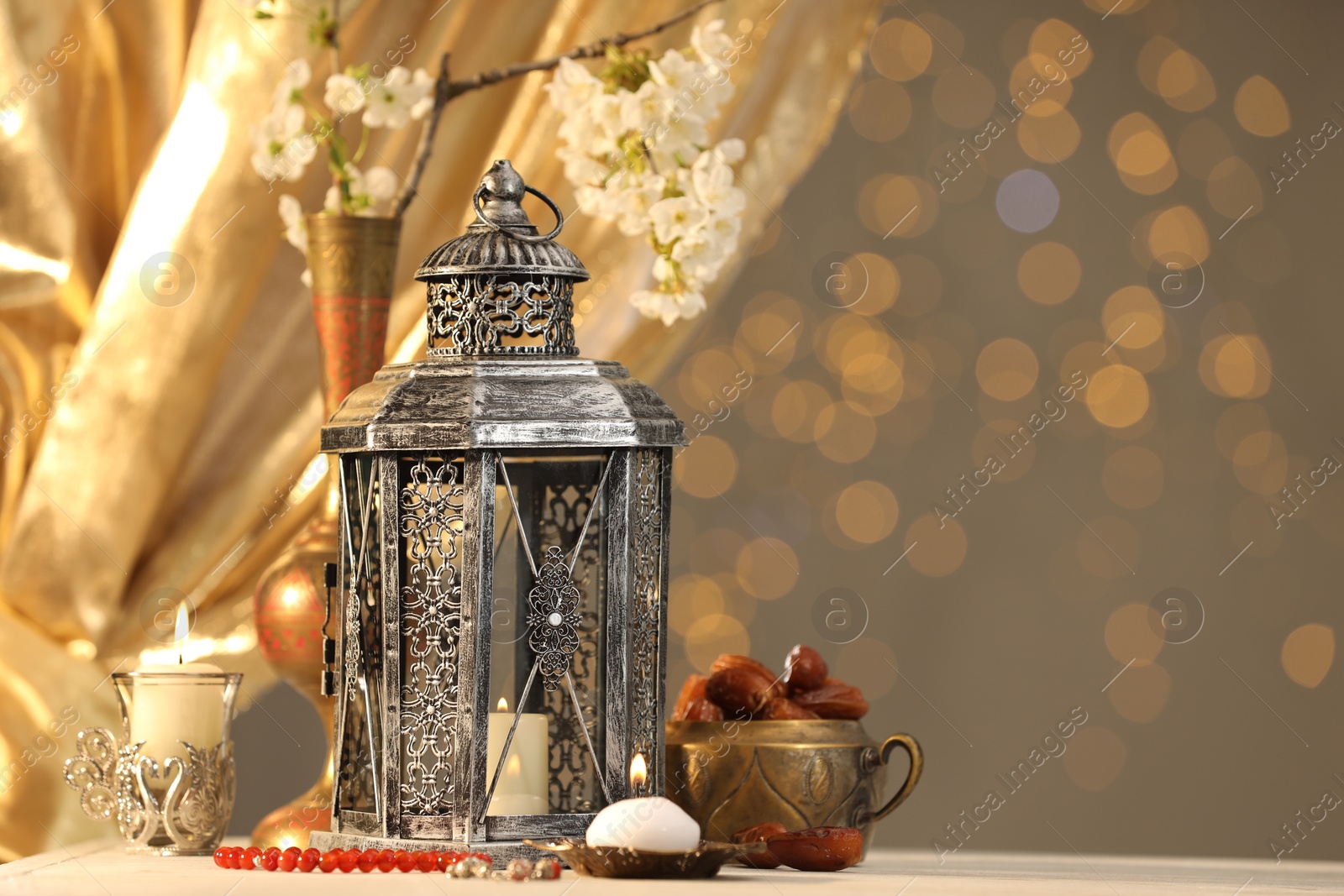 Photo of Arabic lantern, misbaha, candles, dates and flowers on table against blurred lights, space for text