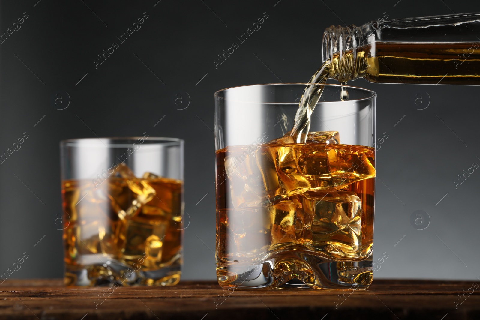 Photo of Pouring whiskey into glass with ice cubes on wooden table against grey background, closeup