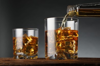 Photo of Pouring whiskey into glass with ice cubes on wooden table against grey background, closeup