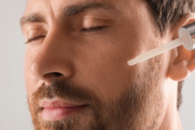 Handsome man applying cosmetic serum onto his face on light grey background, closeup