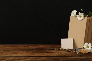 Stationery, glasses and flowers on wooden table near chalkboard, space for text. Teacher's Day