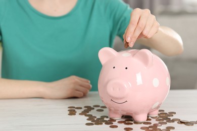 Financial savings. Woman putting coin into piggy bank at white wooden table indoors, closeup