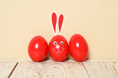 Image of One egg with drawn face and ears as Easter bunny among others on white wooden table against beige background