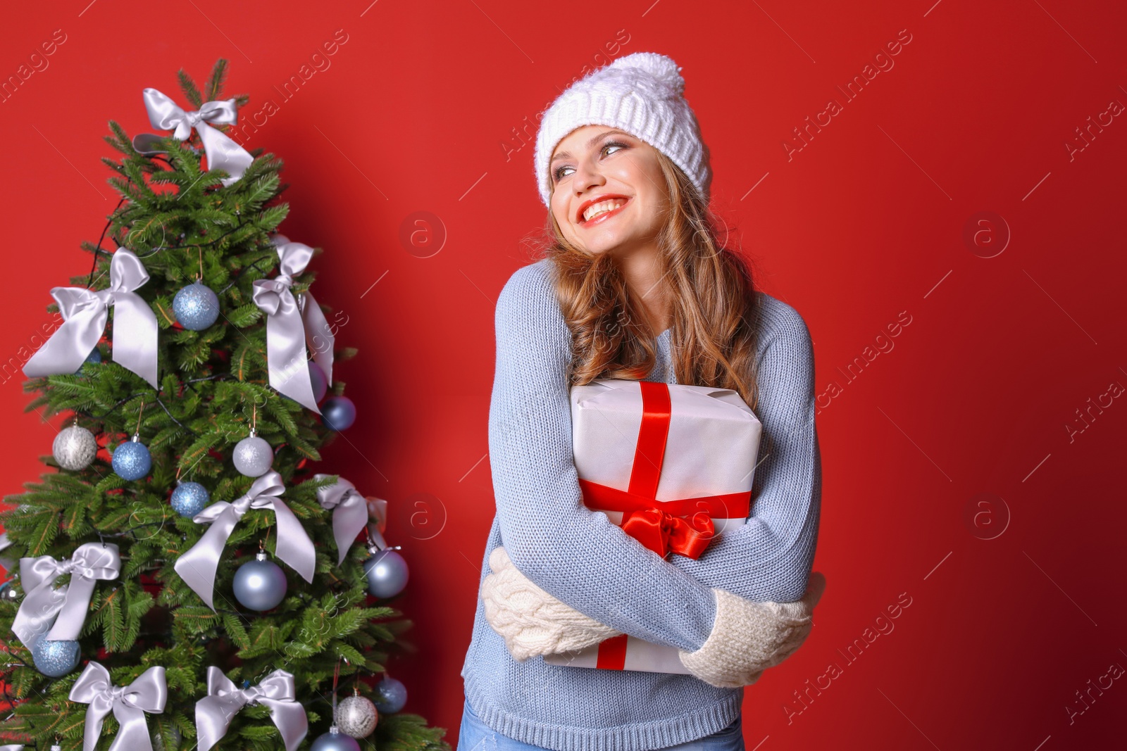 Photo of Beautiful young woman in hat and mittens with gift box near Christmas tree on color background