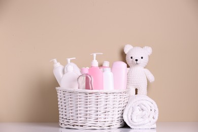 Photo of Wicker basket with baby cosmetic products, bath accessories and knitted toy bear on white table against beige background