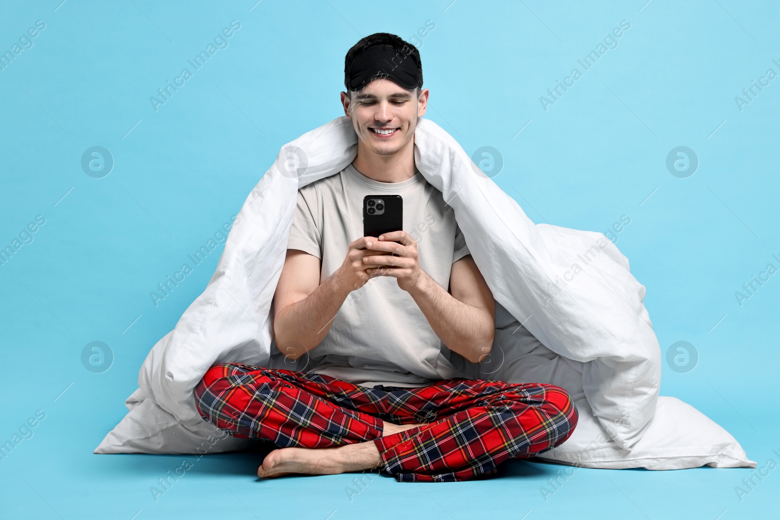 Photo of Happy man in pyjama wrapped in blanket using smartphone on light blue background