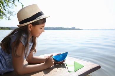 Cute little girl making paper boats on wooden pier near river