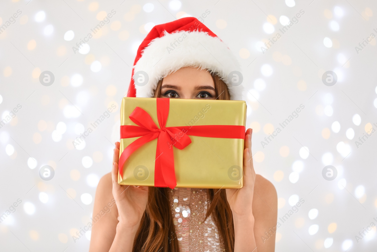 Photo of Happy young woman in Santa hat with gift box against blurred Christmas lights