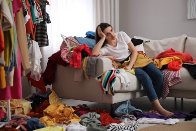 Photo of Upset young woman with lots of clothes on sofa in room. Fast fashion