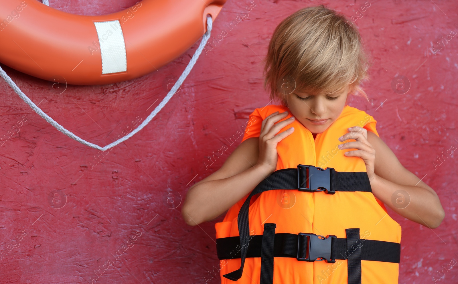 Photo of Little boy putting on orange life vest near red wall with safety ring