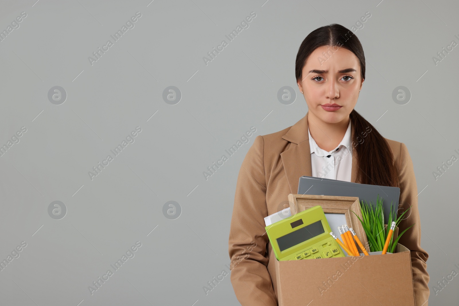 Photo of Unemployment problem. Unhappy woman with box of personal office belongings on grey background, space for text