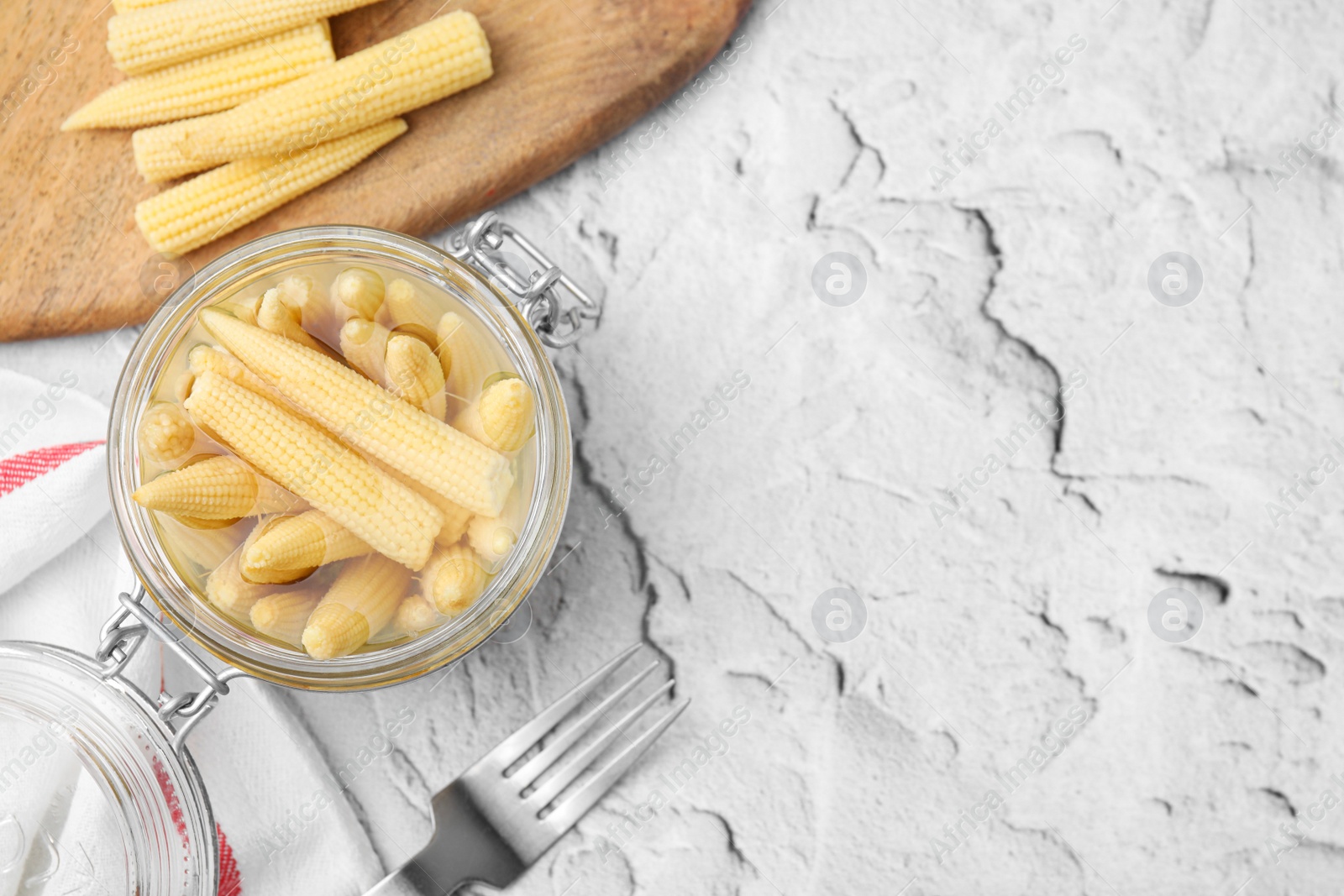 Photo of Jar of pickled baby corn and fork on white textured table, flat lay. Space for text