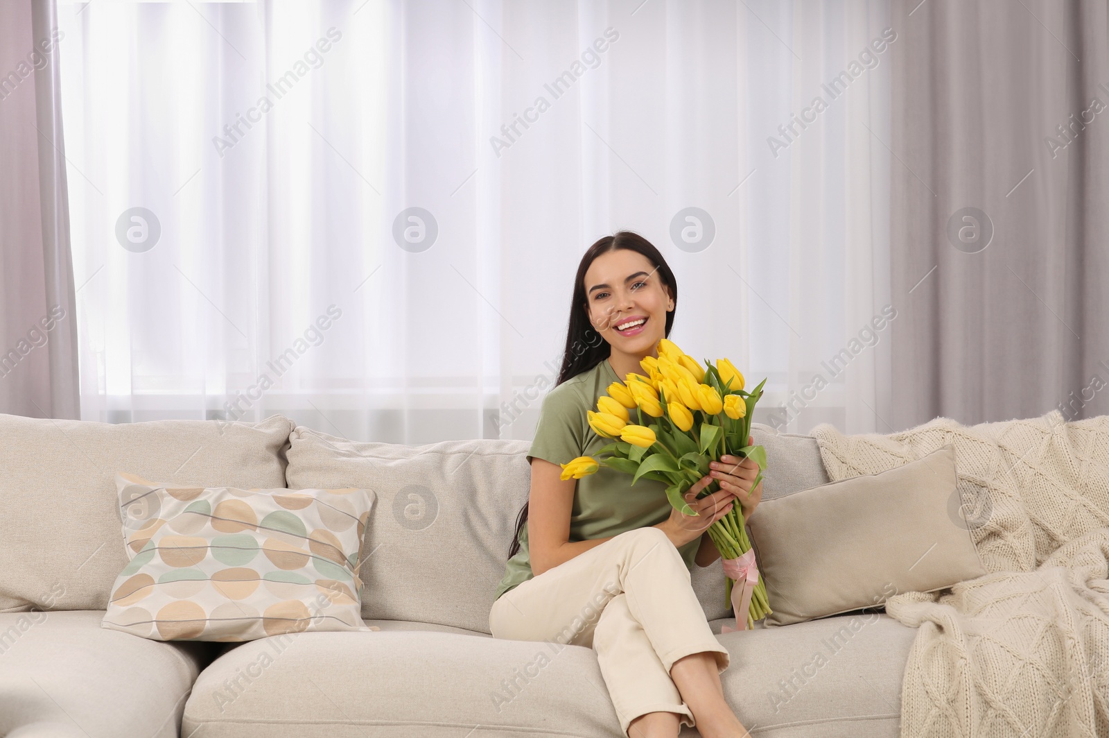 Photo of Happy young woman with bouquet of yellow tulips on couch at home. Space for text