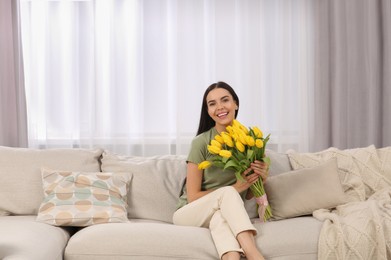 Photo of Happy young woman with bouquet of yellow tulips on couch at home. Space for text