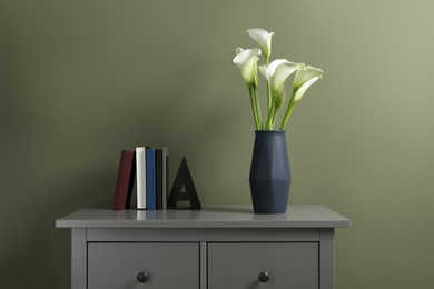 Photo of Beautiful calla lily flowers in vase and books on grey chestdrawers near olive wall
